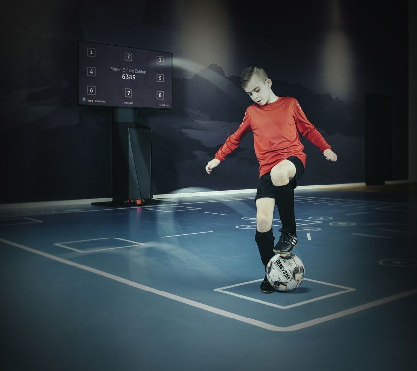 Person spielt Fußball in einer Halle mit einer digitalen Anzeigetafel im Hintergrund